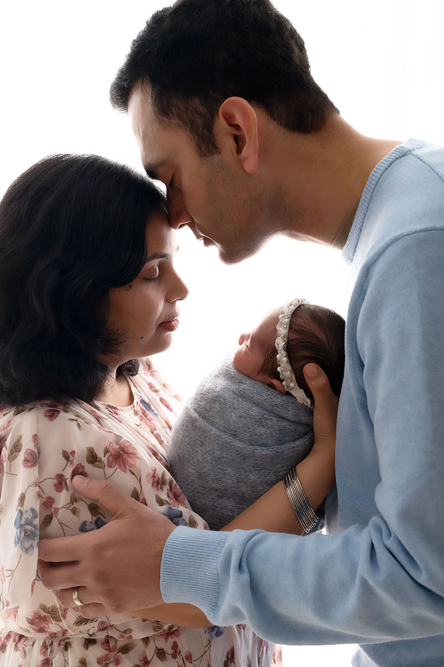 Intimate family moment with parents cradling newborn between them against white backdrop.