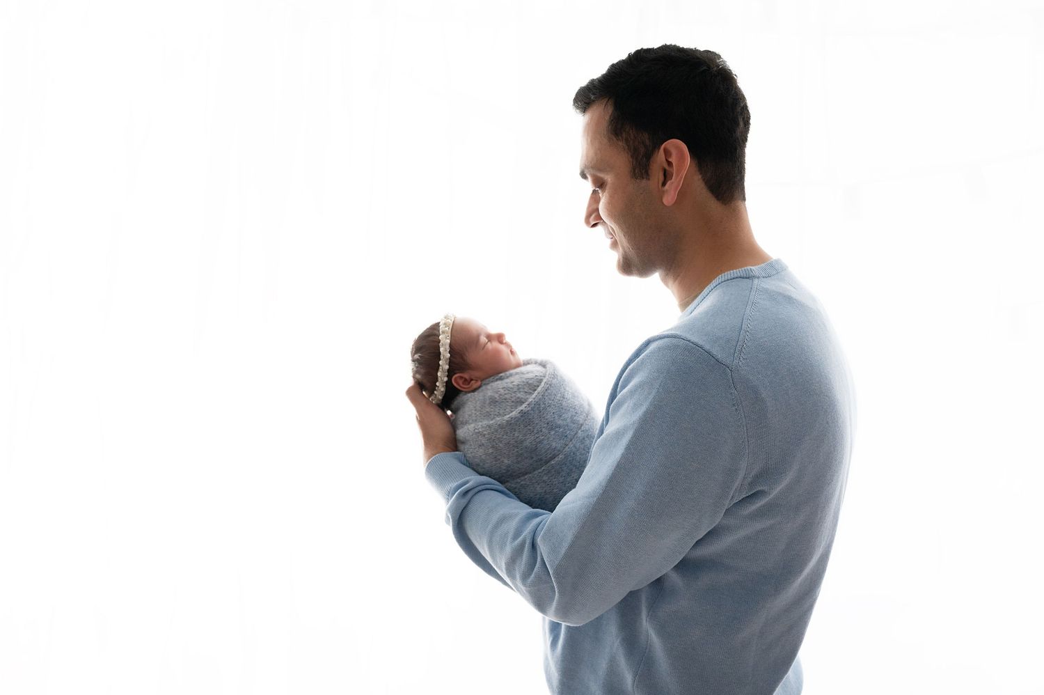 Father in blue sweater tenderly holding swaddled newborn baby against white background.