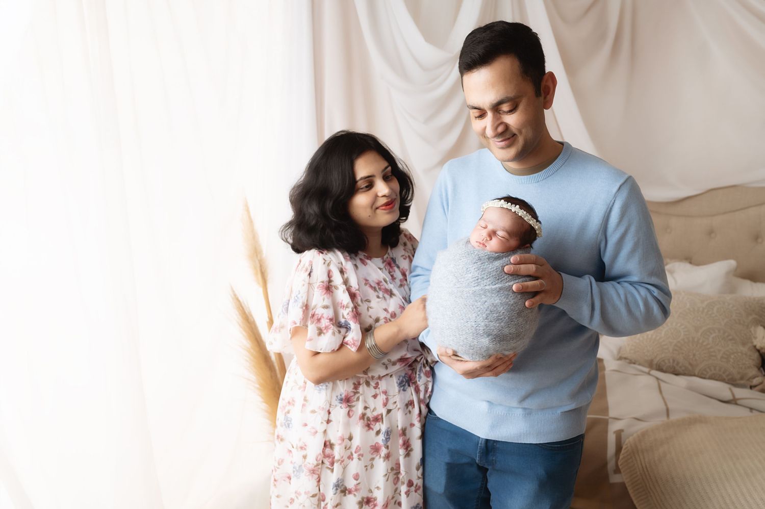 Family standing together against white backdrop with newborn baby wrapped in gray swaddle.