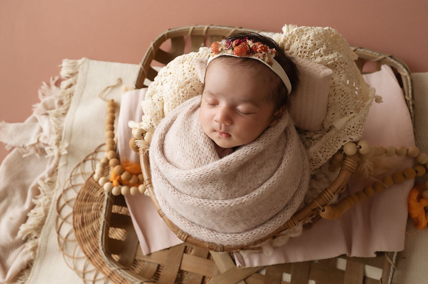 Sleeping newborn in beige wrap and floral headband posed in vintage wooden basket.