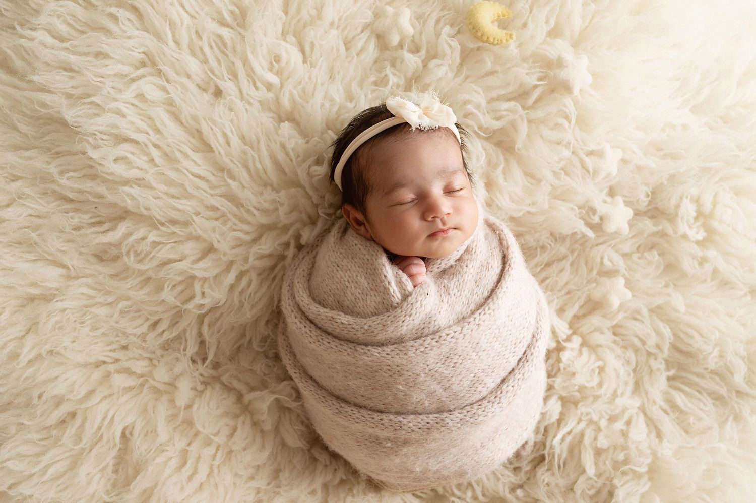 Peaceful sleeping newborn wearing headband and wrapped in beige blanket on fluffy white rug.