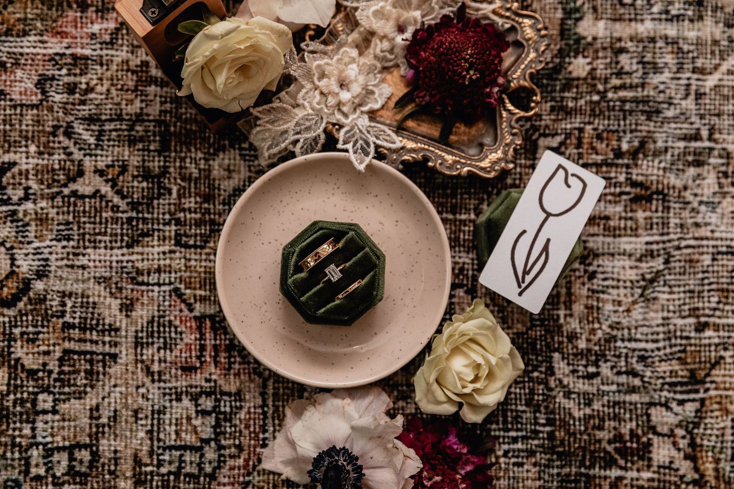 Flat lay of velvet ring box with gold wedding bands, floral lace, florals, and tulip card on vintage rug for romantic wedding details.