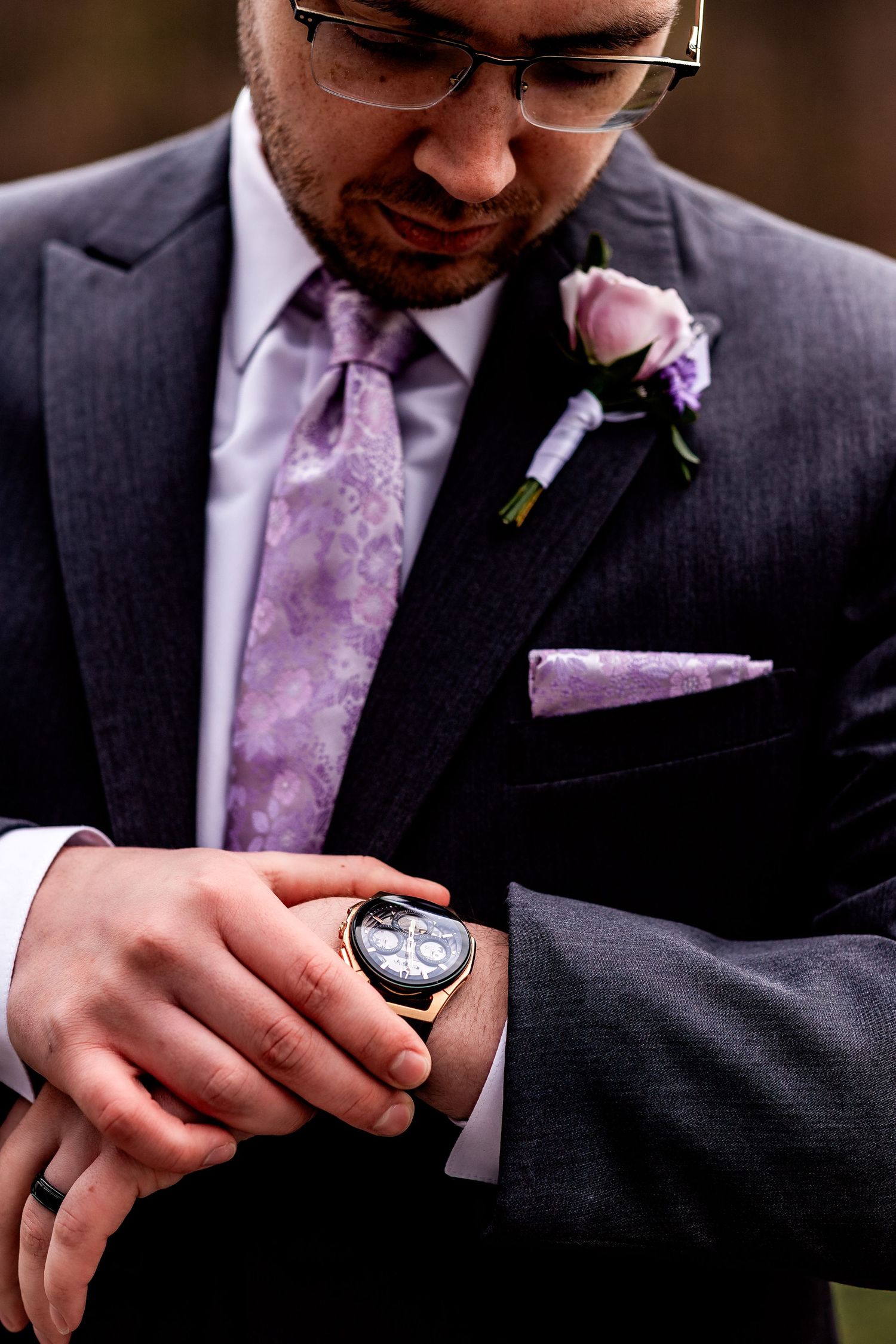 Close-up of groom in charcoal suit with purple tie and boutonniere checking watch on wedding day.