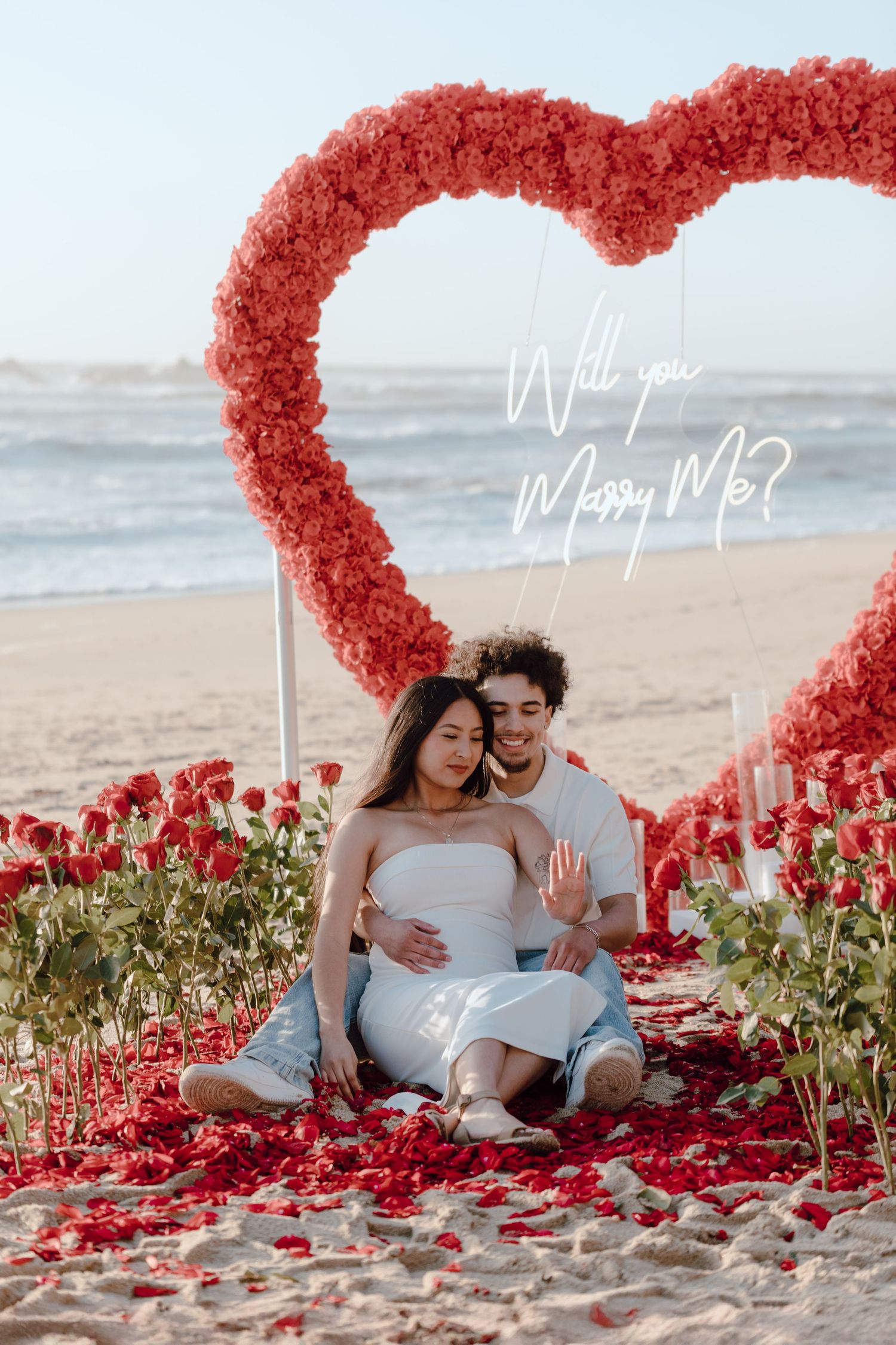 Beach proposal setup with red heart-shaped arch and rose petals on sand.