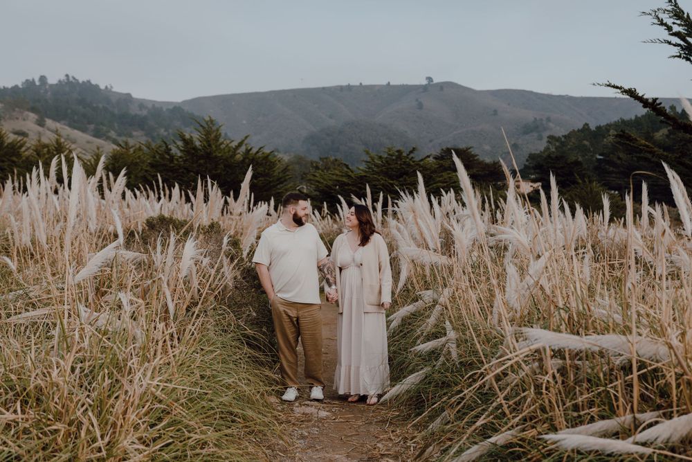 Two people standing on a path surrounded by tall pampas grass against a scenic mountain backdrop.