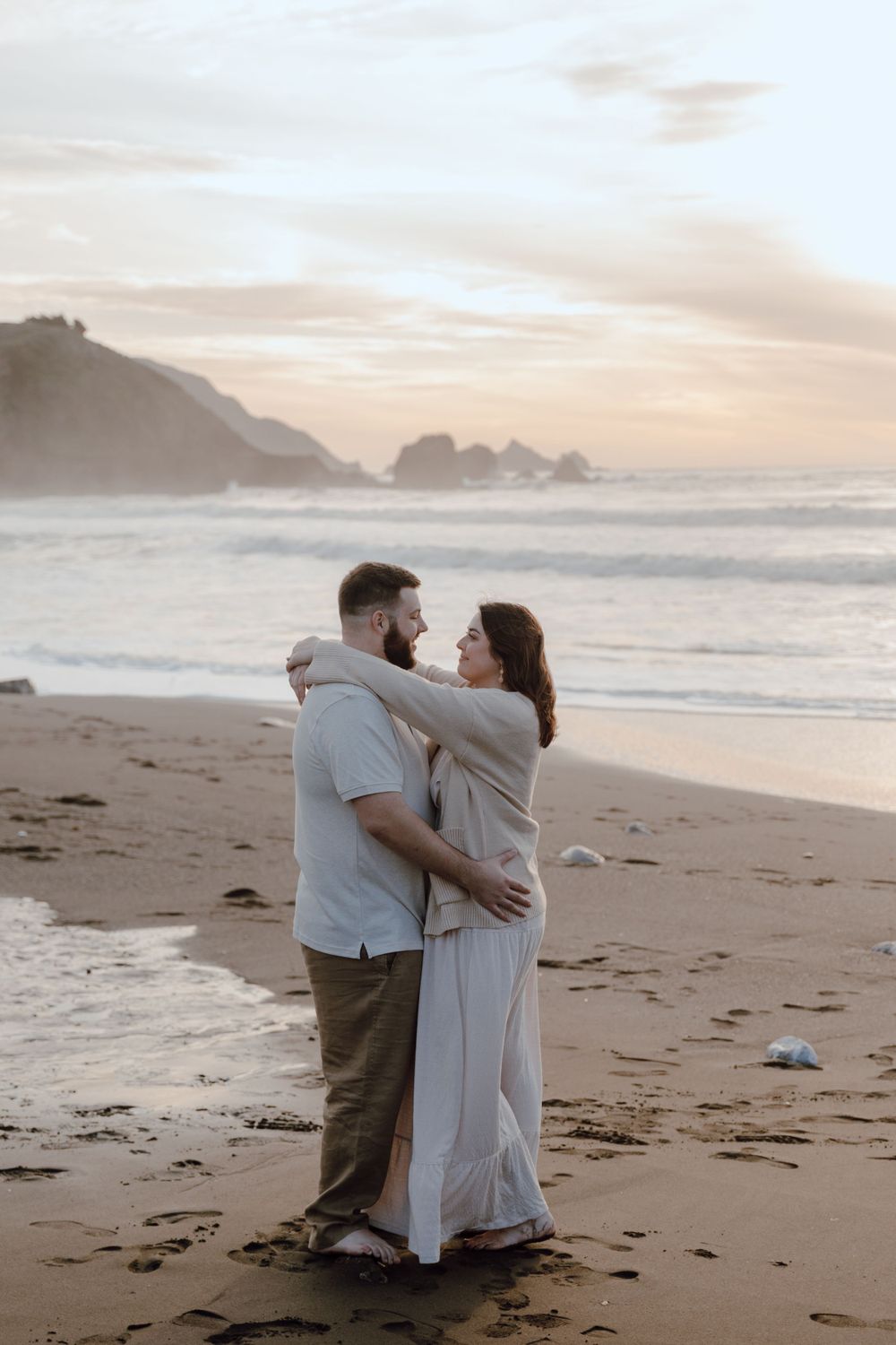 A couple embraces on a misty beach at sunset with dramatic coastal cliffs and crashing waves in the background.