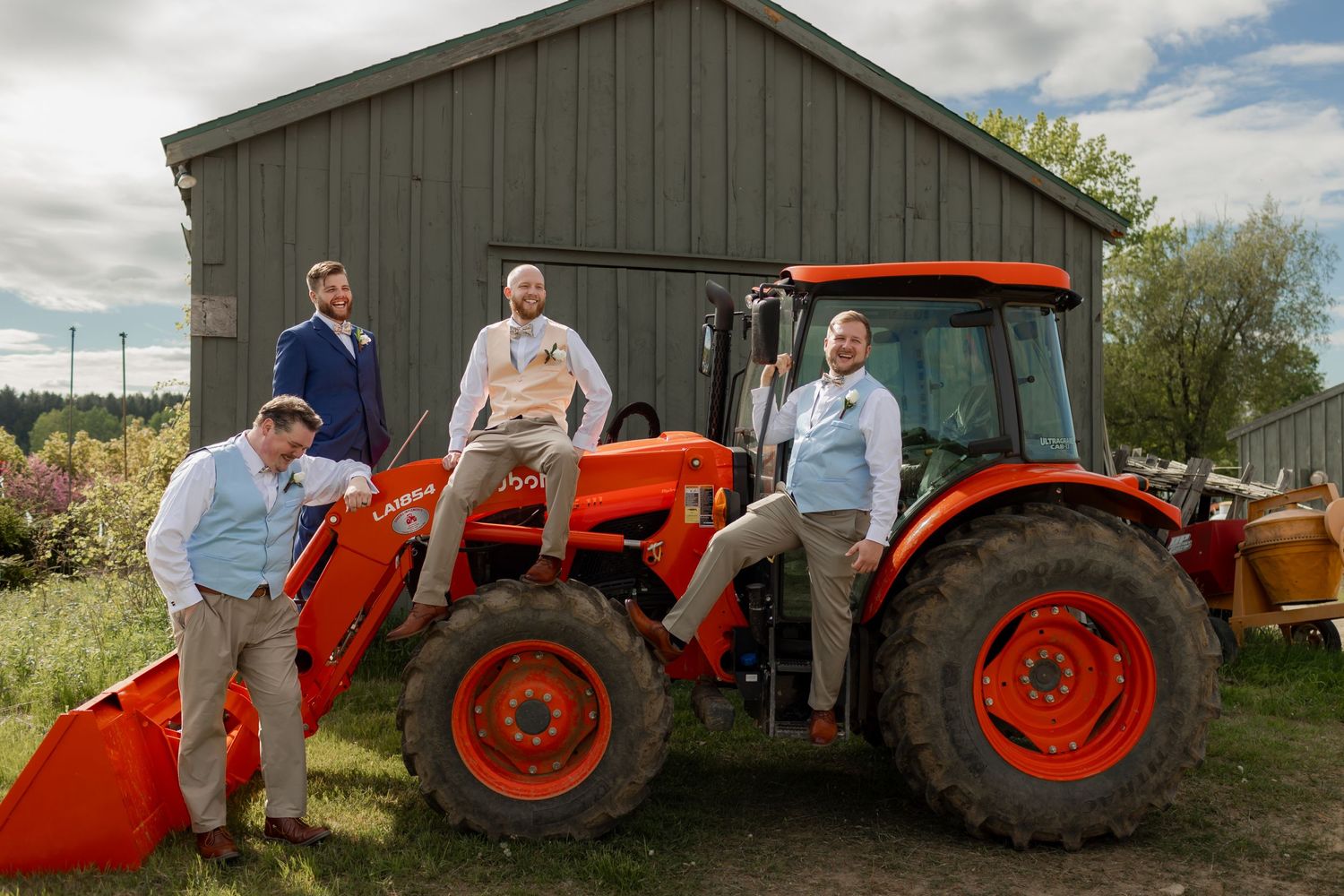 An orange Kubota tractor with a front loader bucket stands in front of a gray barn on a sunny day.