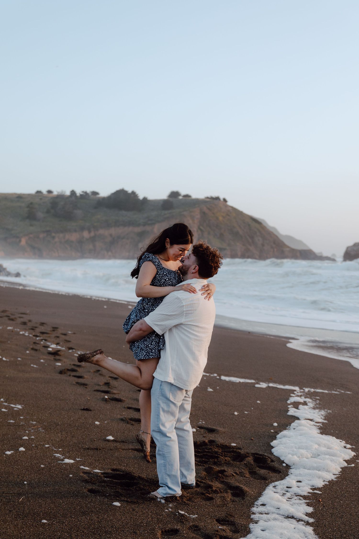 Couple dancing together on foggy coastal cliffs overlooking a dramatic rocky shoreline and ocean.