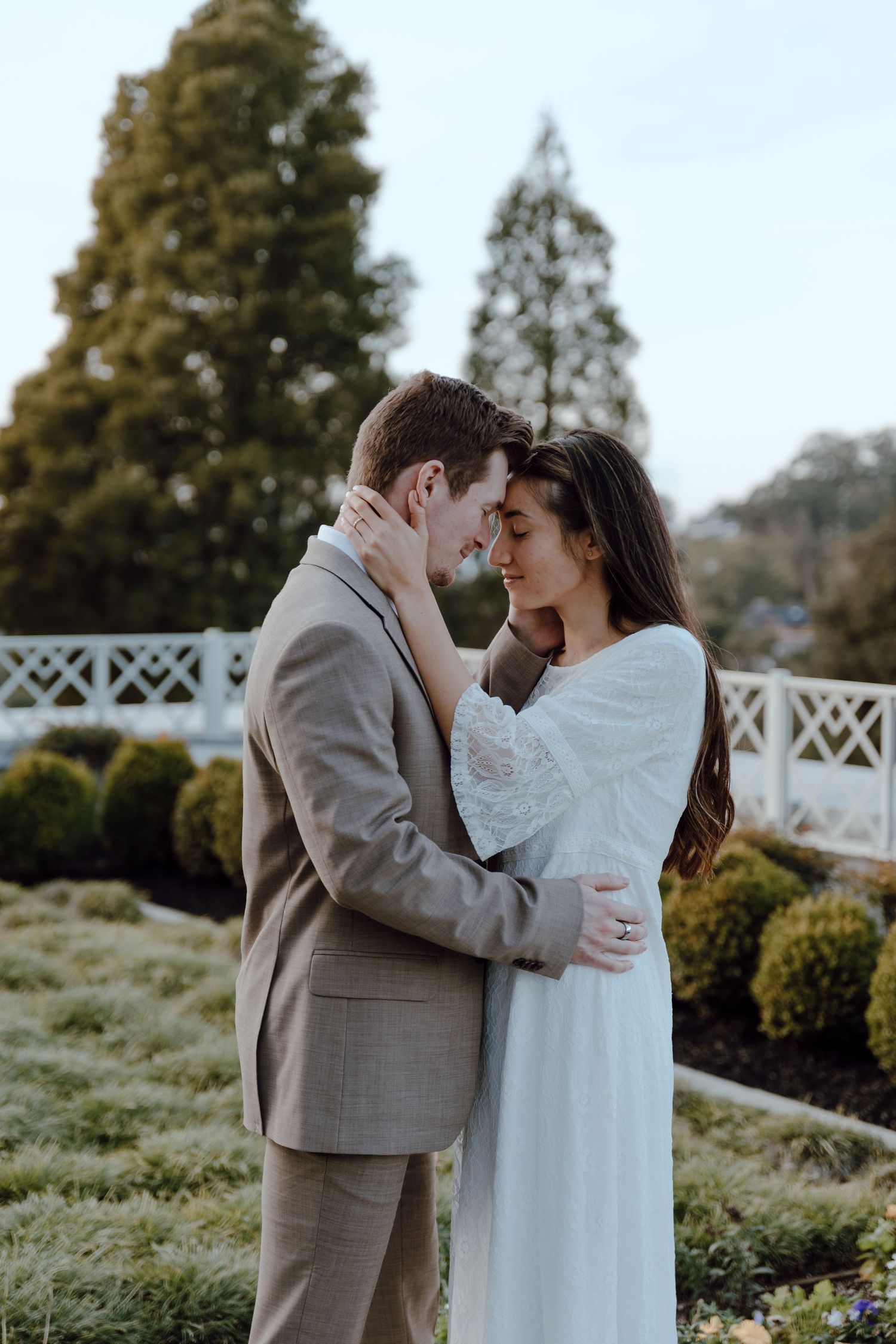 A couple shares a tender moment outdoors with green foliage and warm sunset light in the background.