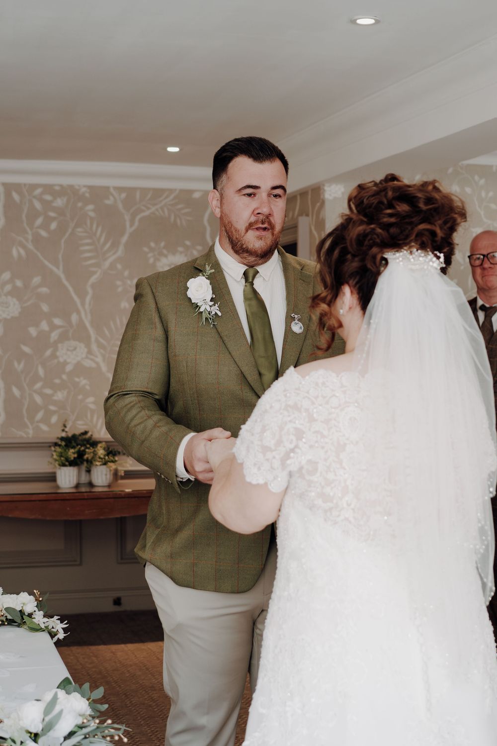A couple exchanges wedding vows in an elegant indoor venue with floral wallpaper and recessed lighting.