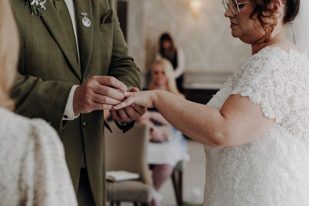Close-up view of hands exchanging wedding rings during an indoor ceremony with green suit and white lace dress details.