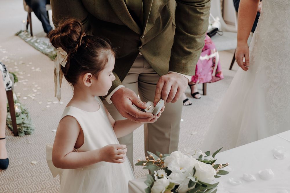 Small child in a white dress stands near floral arrangements during wedding ceremony.