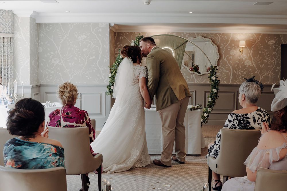 A couple shares a first kiss during an intimate indoor wedding ceremony beneath a floral arch with seated guests watching.