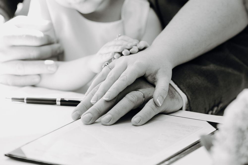 Two hands touching tenderly during a document signing or consultation meeting with pen and paper visible on desk.