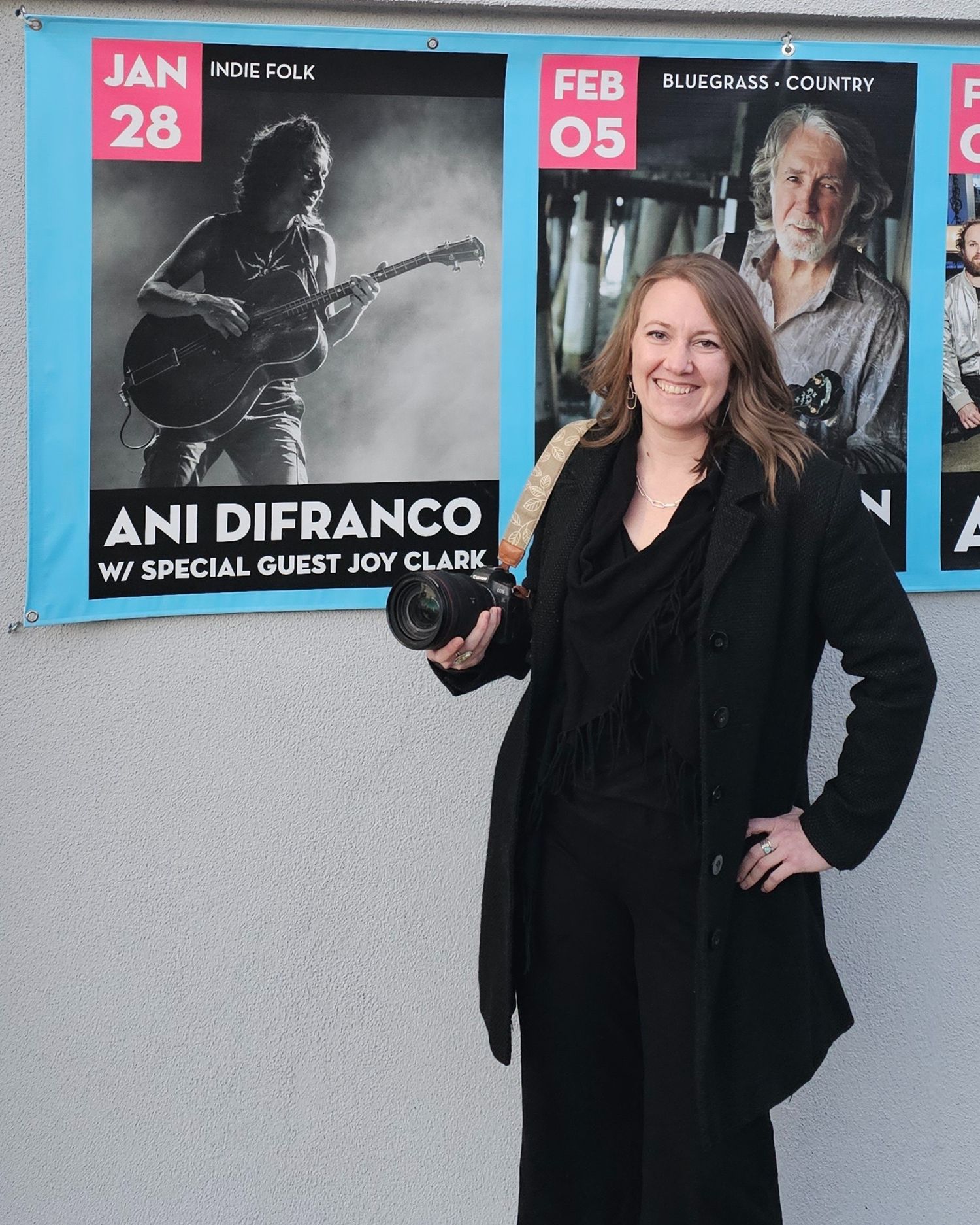 A person in a black coat stands next to concert promotional posters on a white wall.