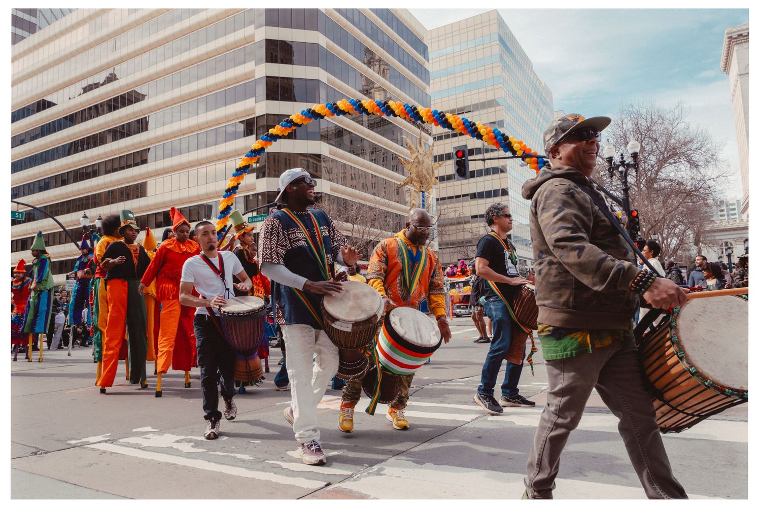 A parade of musicians in colorful attire march through city streets with drums and instruments during a festive celebration.