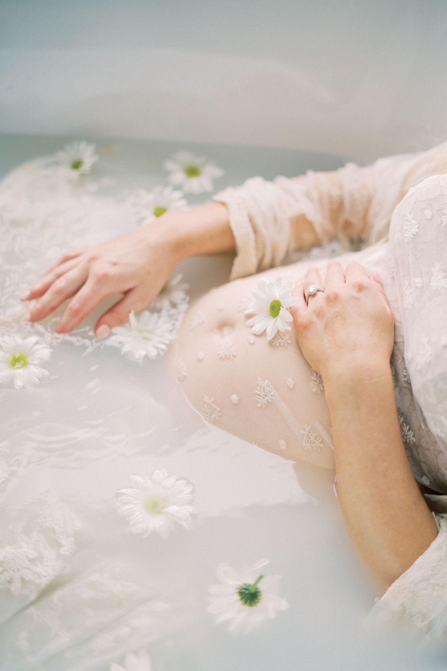 Hands resting on delicate white fabric surrounded by scattered daisies in a dreamy milk bath composition.