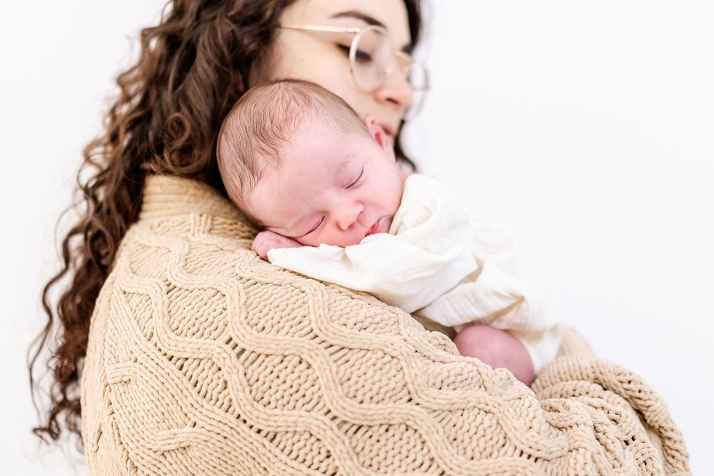 A newborn baby sleeps peacefully against a shoulder wrapped in a cozy beige cable knit sweater.