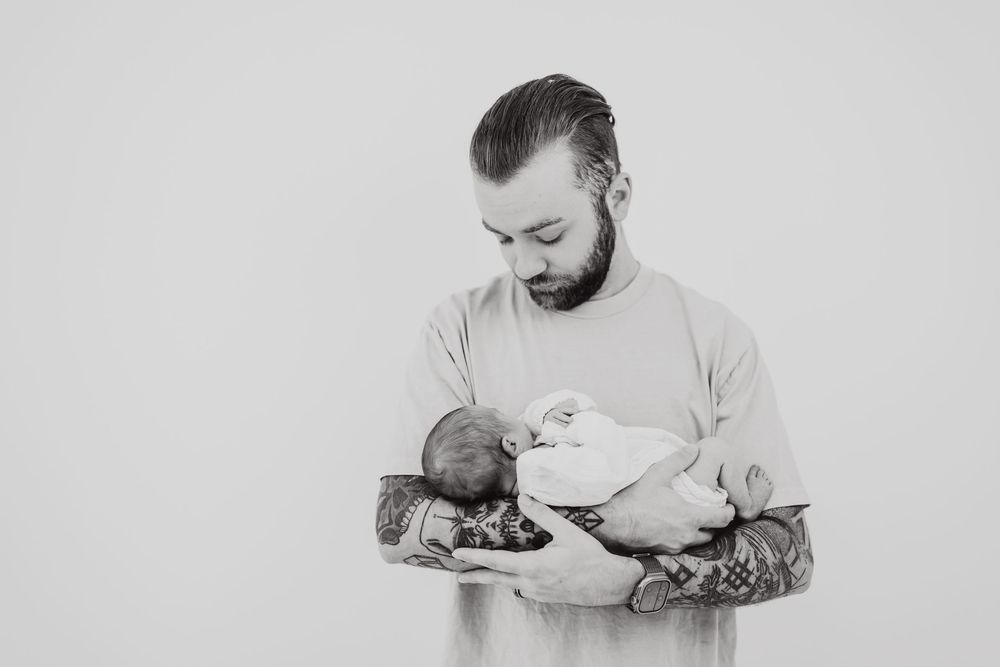 A tattooed person wearing light colored clothing tenderly holds and gazes at a newborn baby in black and white.