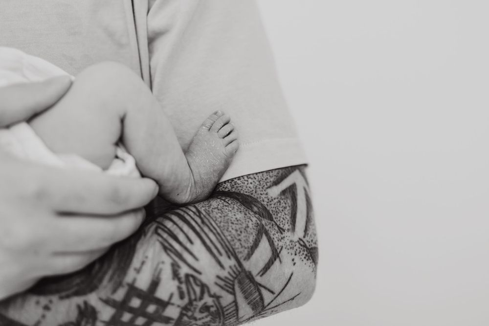 Black and white image of tattooed arms cradling a newborn baby's feet.