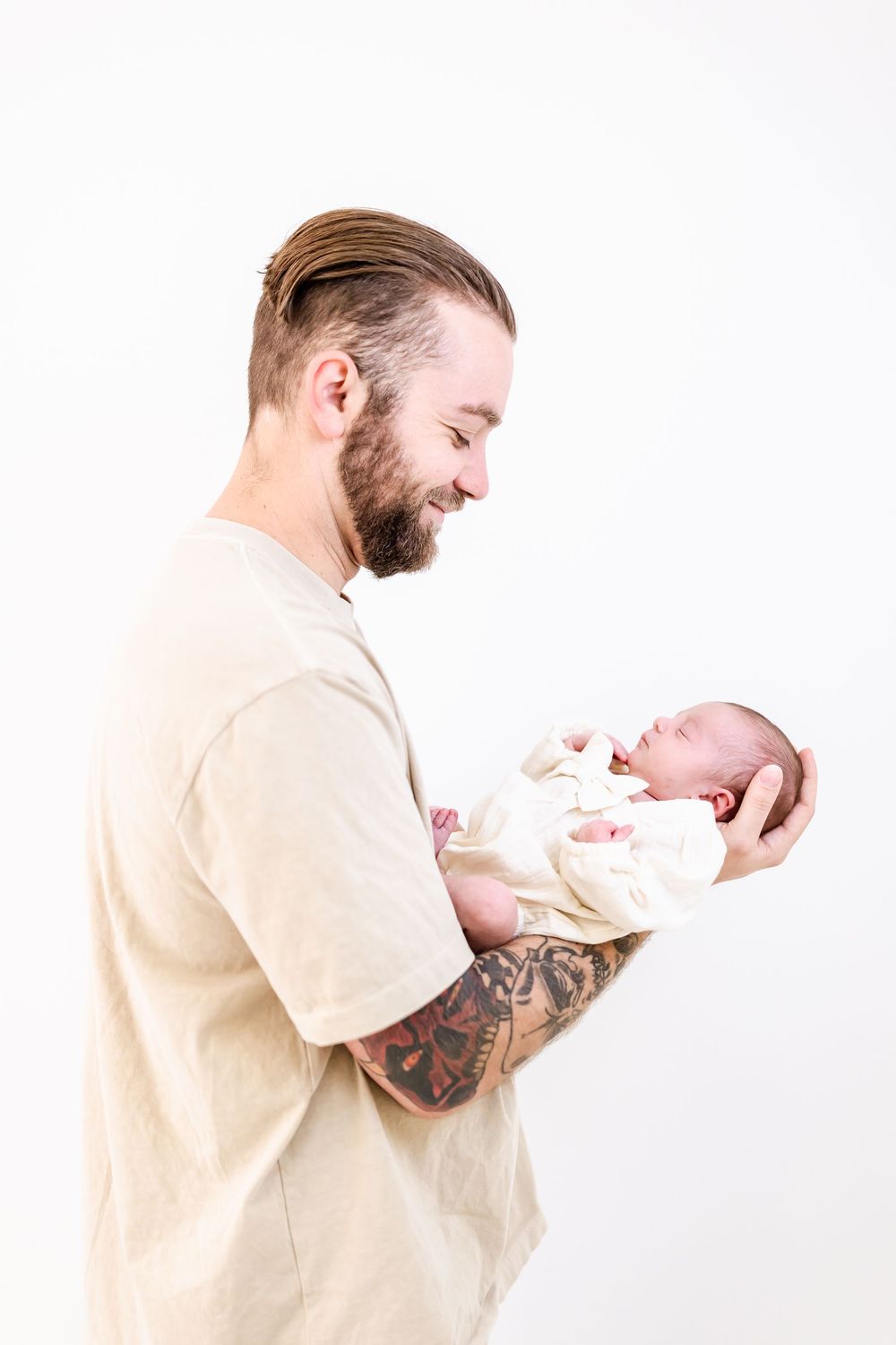 A bearded man in a beige shirt with tattooed arms gazes lovingly at a newborn baby bundled in white.