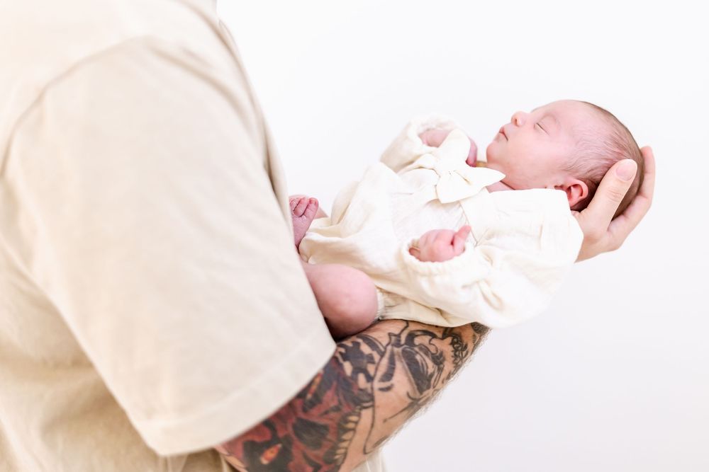 Parent with visible tattoos holds a newborn baby wearing a white outfit against a light background.