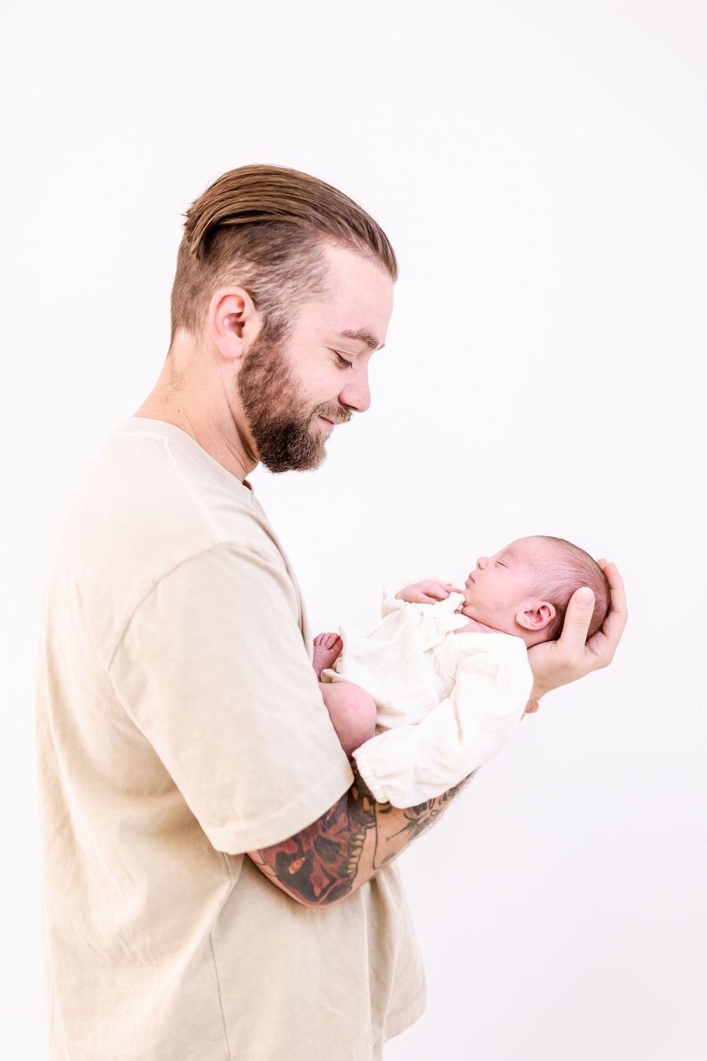 A person in a beige shirt holds and gazes lovingly at a newborn infant in a white outfit against a light background.
