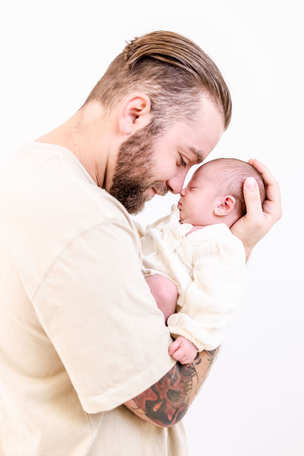 Adult with tattoos and beard tenderly shares a close intimate moment with newborn baby in cream colored clothing against white background.