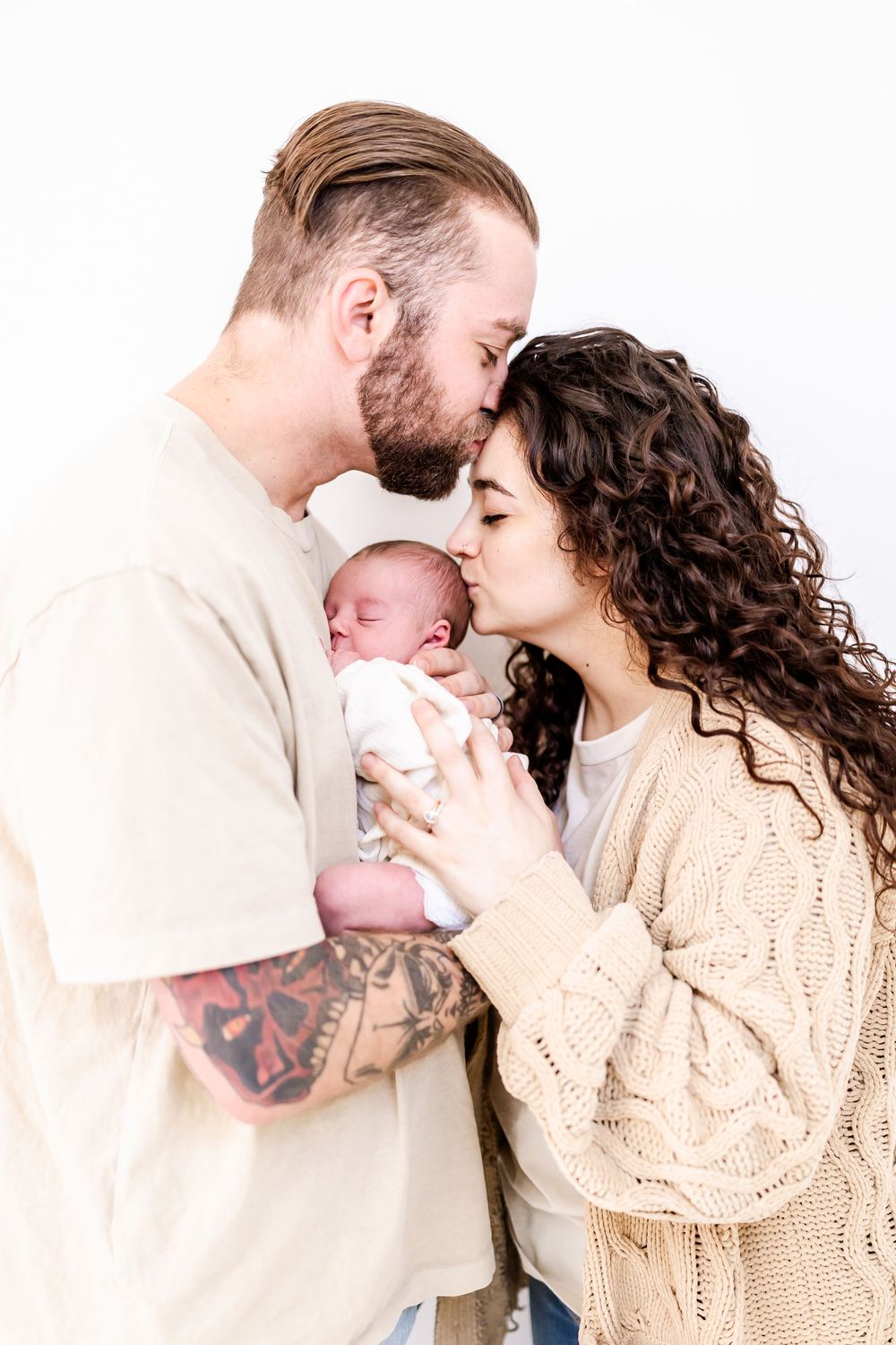 A couple in neutral tones share a tender moment with their newborn baby against a white background.
