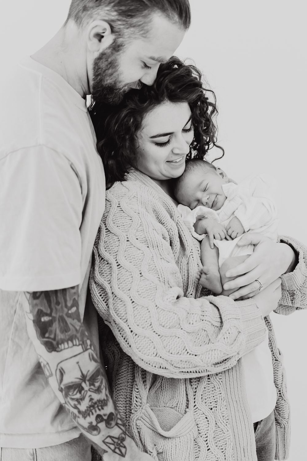Sweet black and white family portrait showing tender moment with newborn baby as parents embrace in knit sweater.