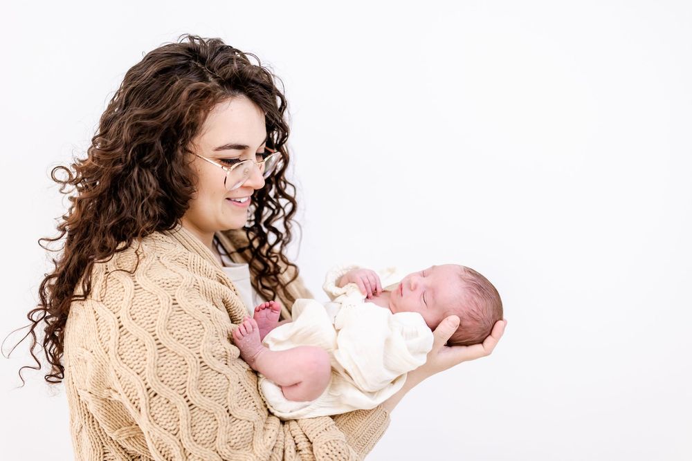 Someone wearing a beige cable knit sweater holds a newborn baby swaddled in white against a bright white background.