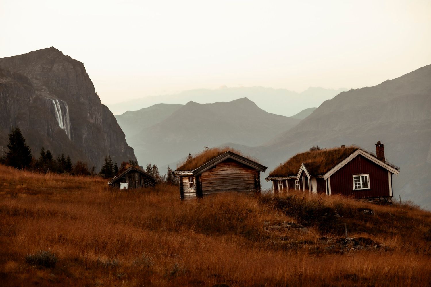 Rustic wooden cabins nestled on a golden hillside with dramatic mountain peaks in the background at sunset.