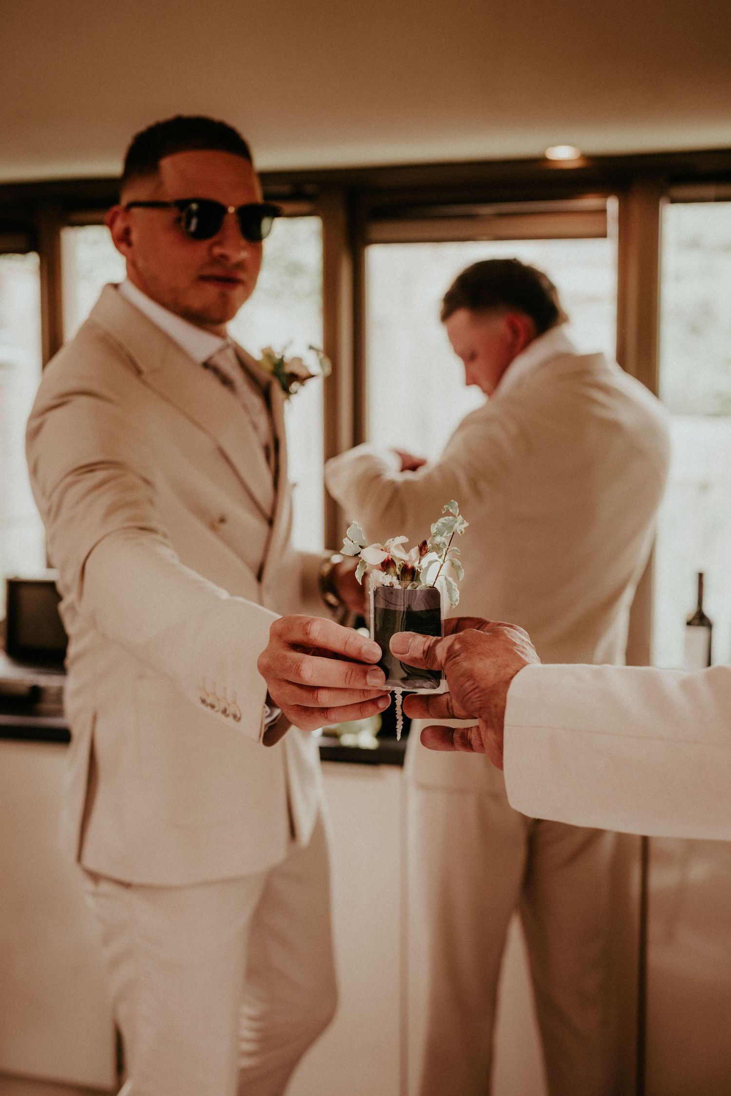 A person in a cream-colored suit and sunglasses holds a glass during what appears to be a wedding celebration.