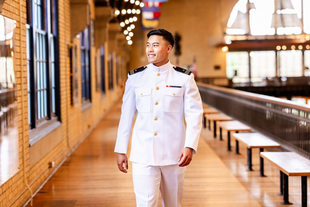 Naval officer stands in crisp white uniform inside rustic venue with wooden benches and hanging lights.