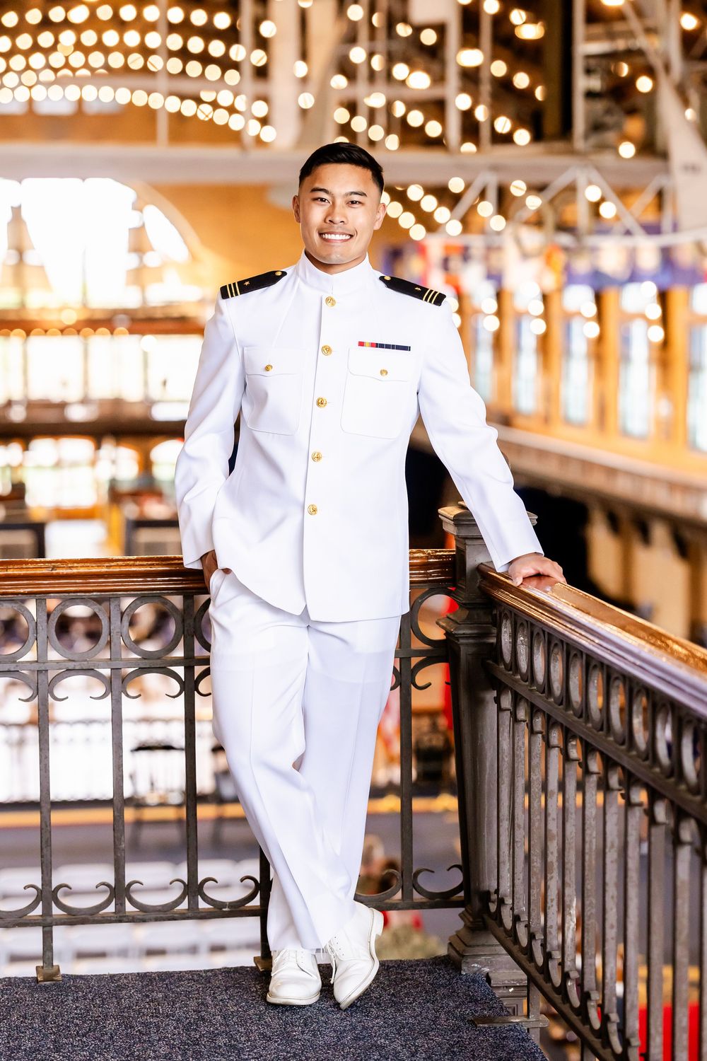 Military officer in white dress uniform posing on illuminated walkway with festive lights in background.