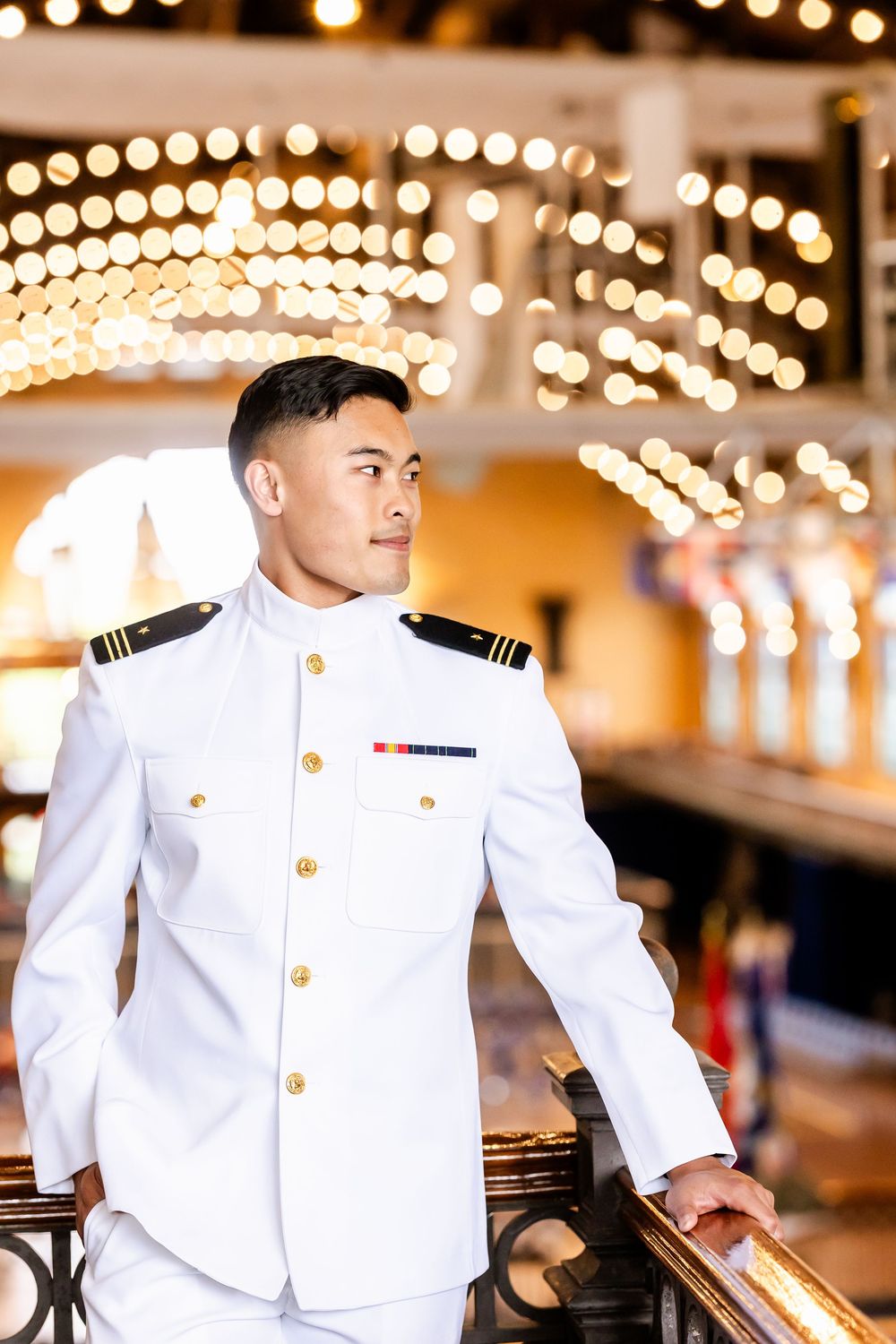 Naval officer in white dress uniform stands on deck with glowing string lights creating a magical bokeh background.