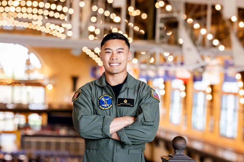 Military service member in flight suit stands confidently in front of decorative lights at indoor venue.