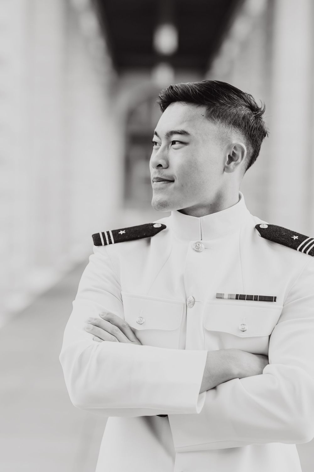 Naval officer in white dress uniform stands with arms crossed in black and white portrait photograph.