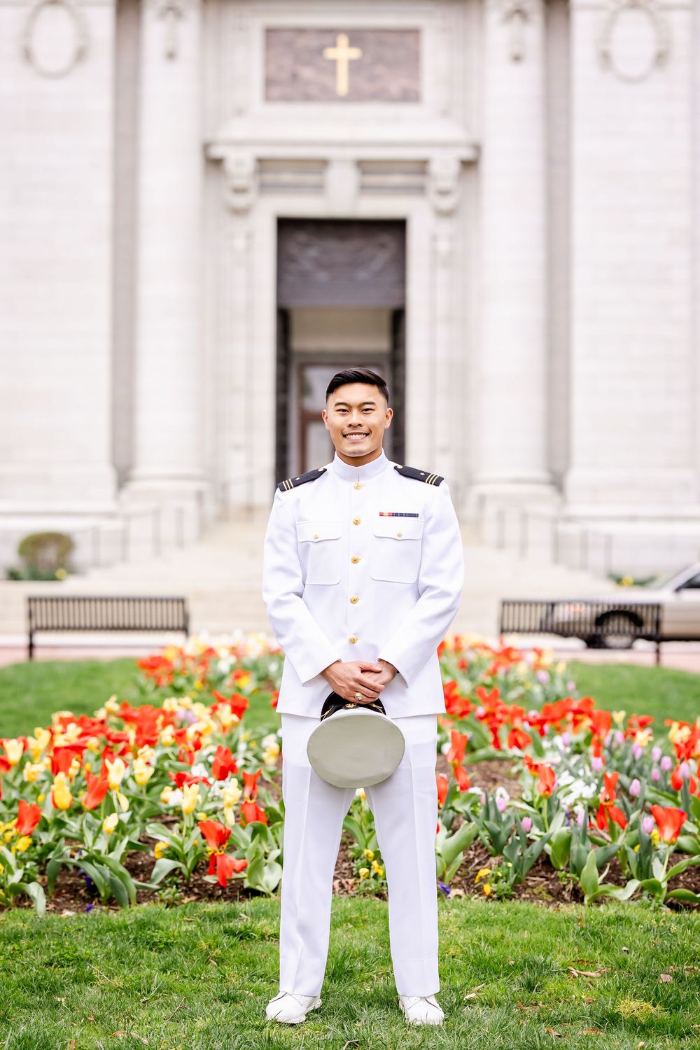Military service member standing in formal white uniform in front of church entrance with blooming tulip garden.