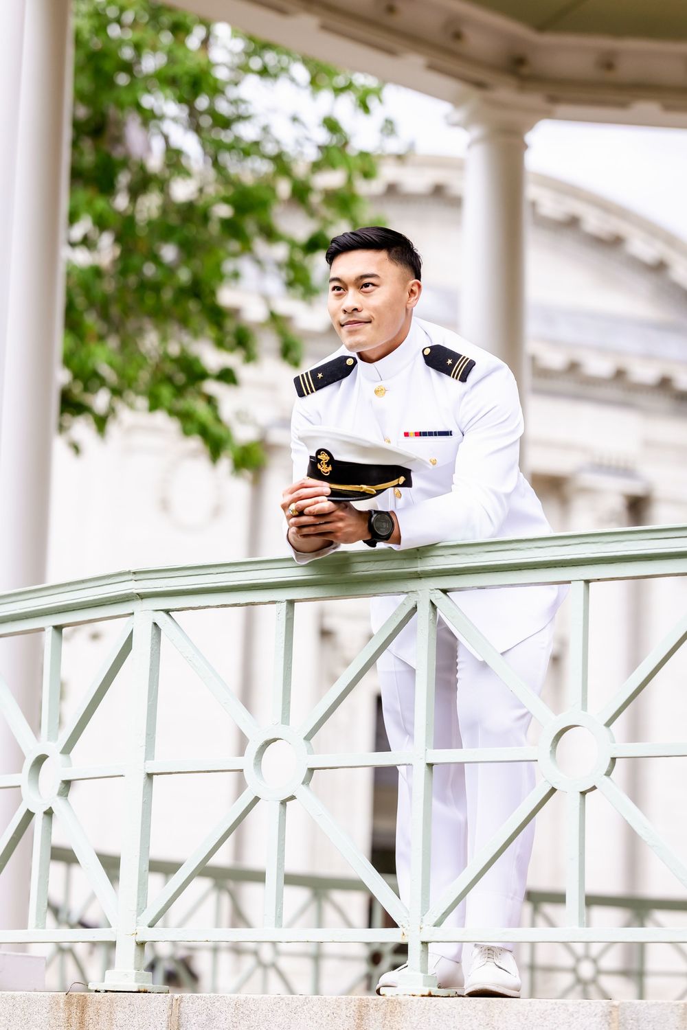 A naval officer in white dress uniform stands on a balcony railing with classical architectural columns in the background.