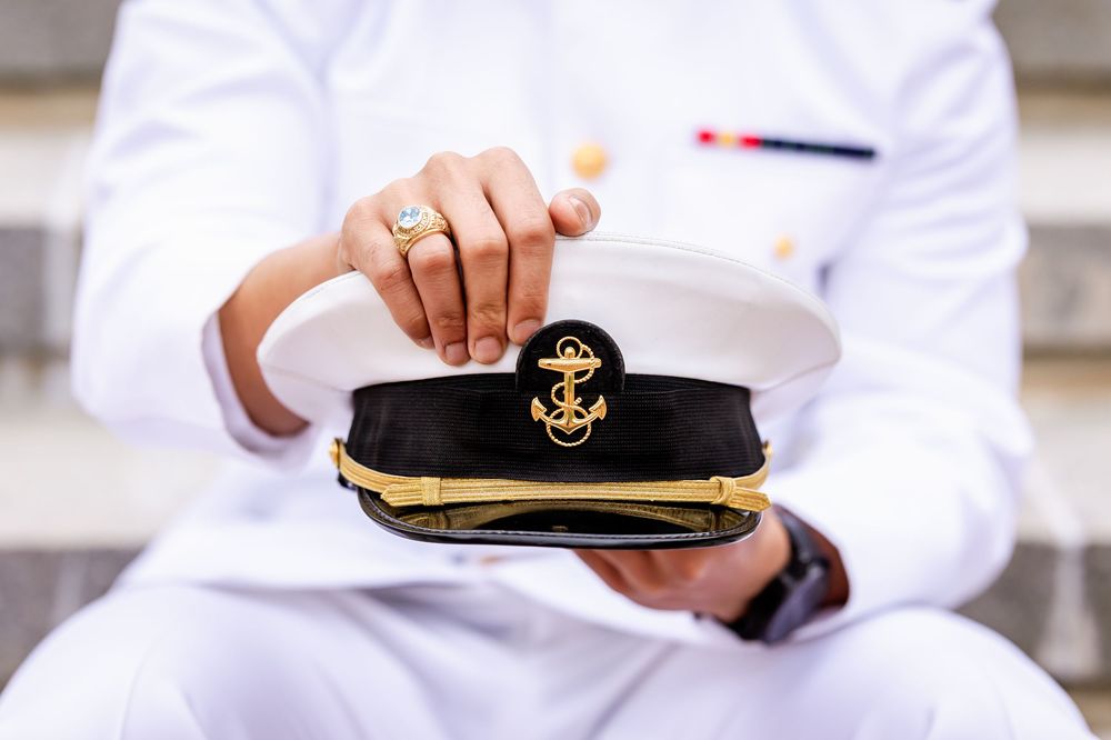 Hands in white uniform hold a naval officer's cap with gold braid and anchor emblem against blurred background.