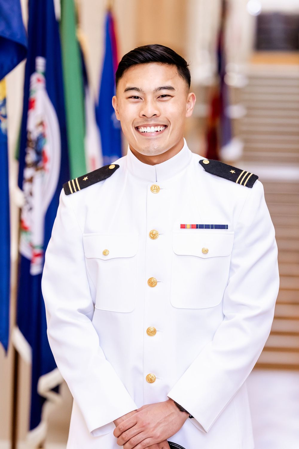Naval officer in white dress uniform with gold buttons stands proudly in front of state flags.