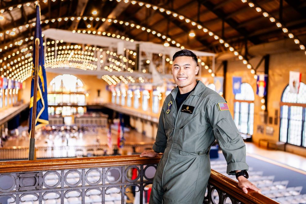 Military service member in flight suit poses inside a historic venue with decorative string lights overhead.