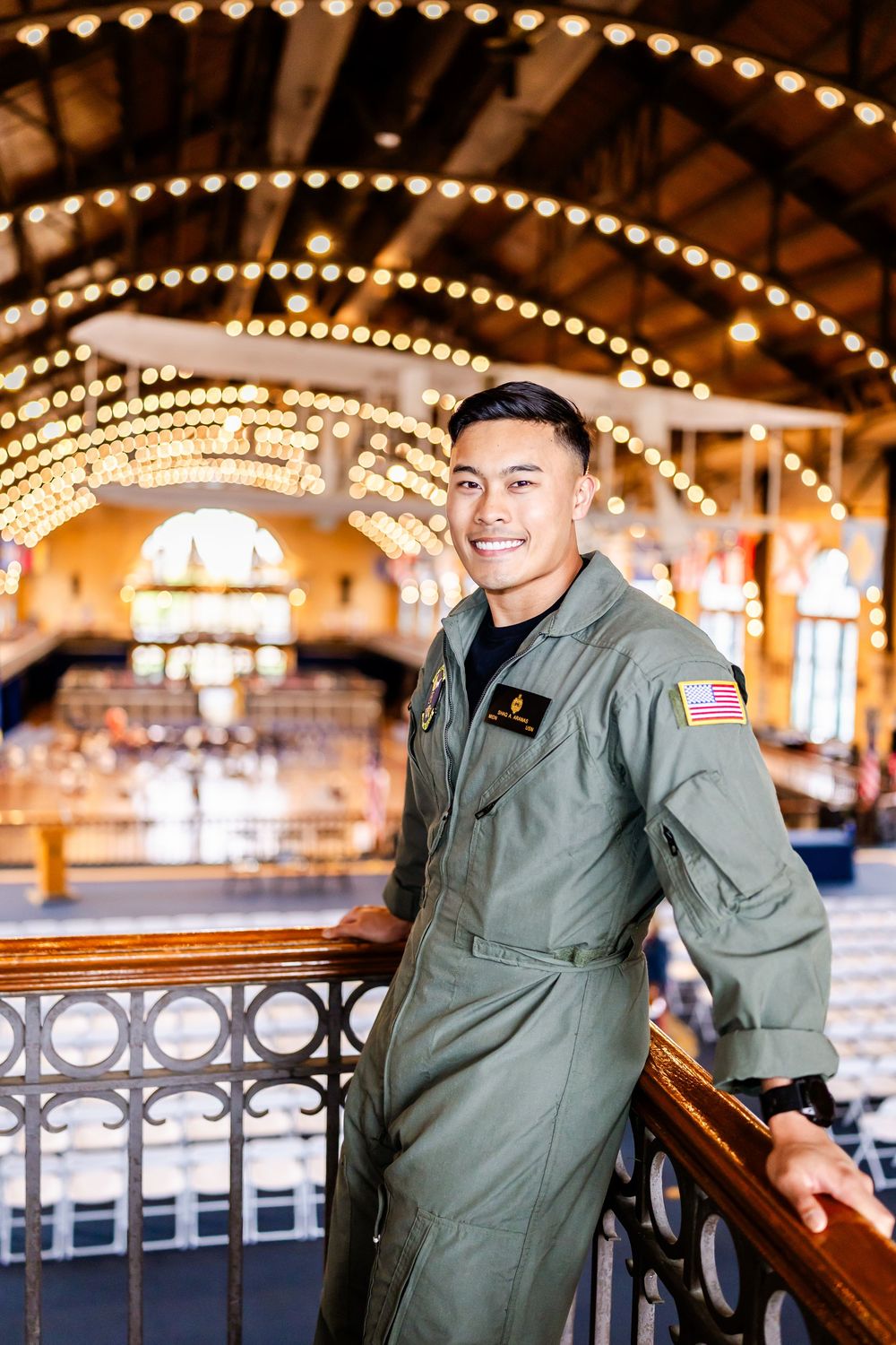 Military service member in flight suit stands in elegant venue with arched ceiling decorated with string lights and chandeliers.