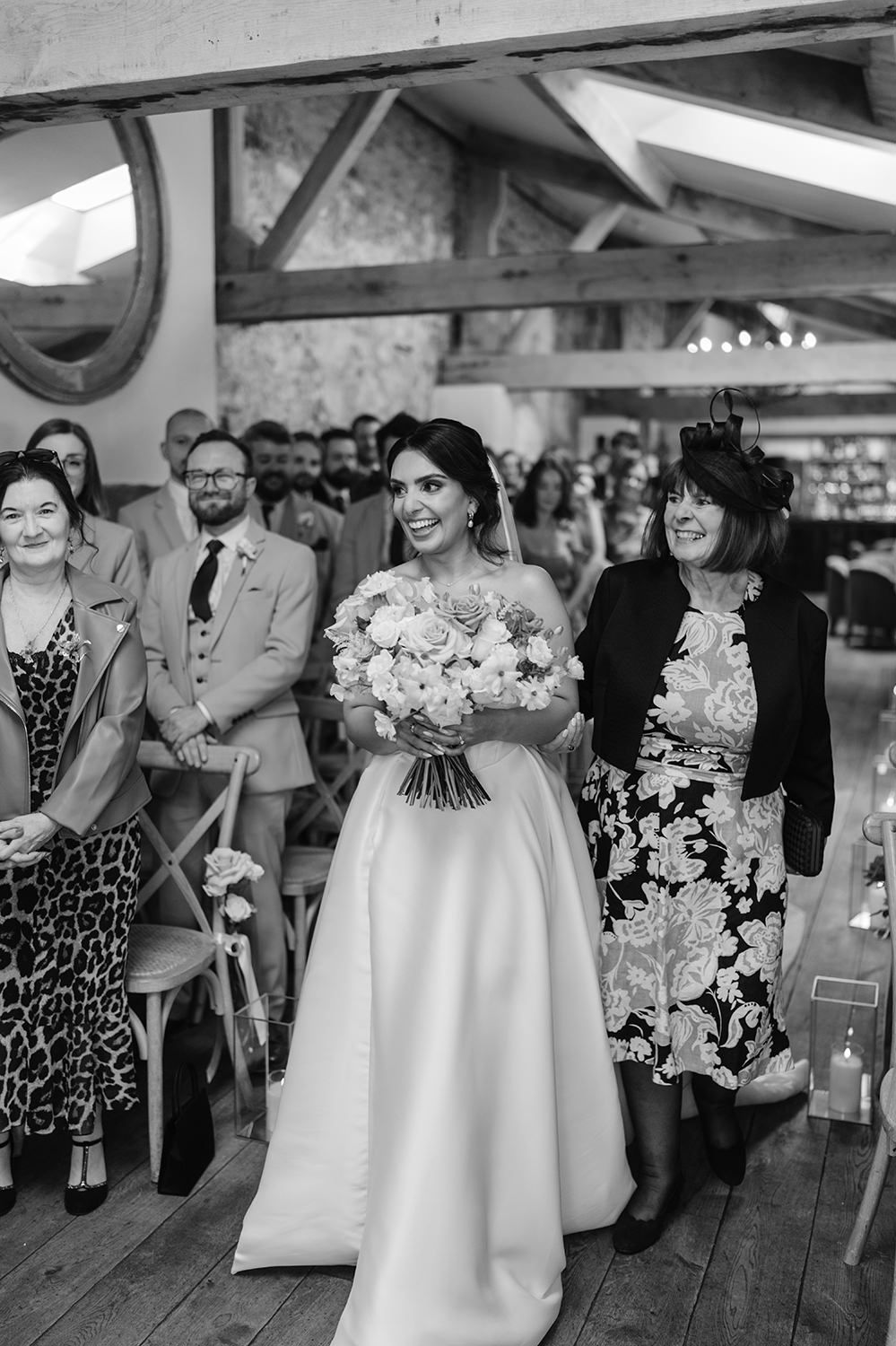 A black and white wedding photo showing wedding guests and a bride in a classic white gown holding a floral bouquet.