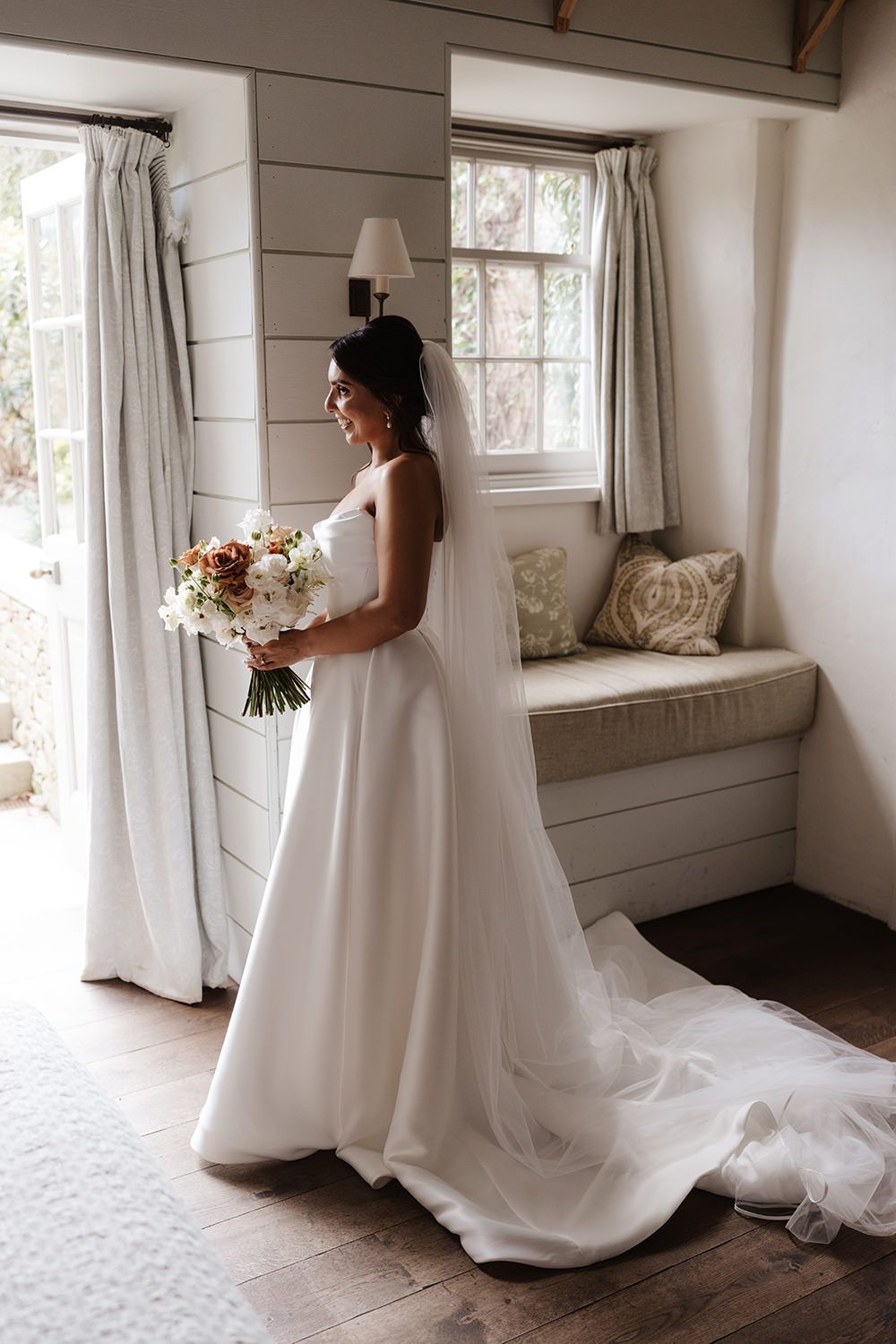 An elegant white wedding dress with a flowing train is shown in a sun-lit room with shiplap walls and a built-in bench.