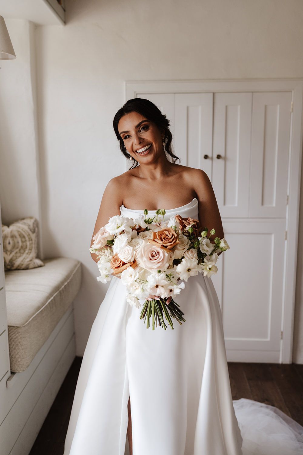 A bride in a classic white strapless wedding gown holds a romantic bouquet of blush roses and white flowers against white doors.
