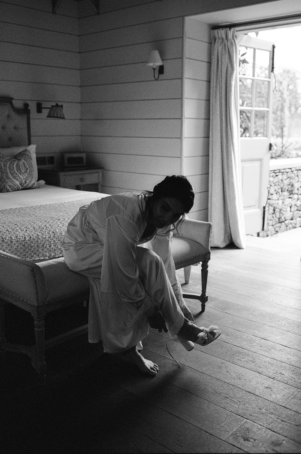 Black and white photo of someone sitting on edge of bed in rustic room with wooden floors and walls near bright window.