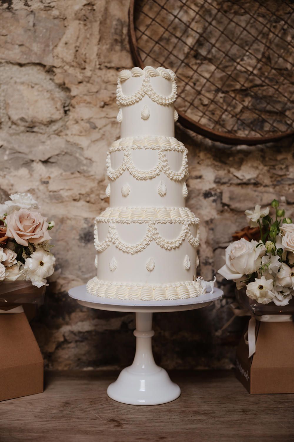 Elegant white tiered wedding cake with pearl swag details on a pedestal stand, flanked by blush and white floral arrangements.