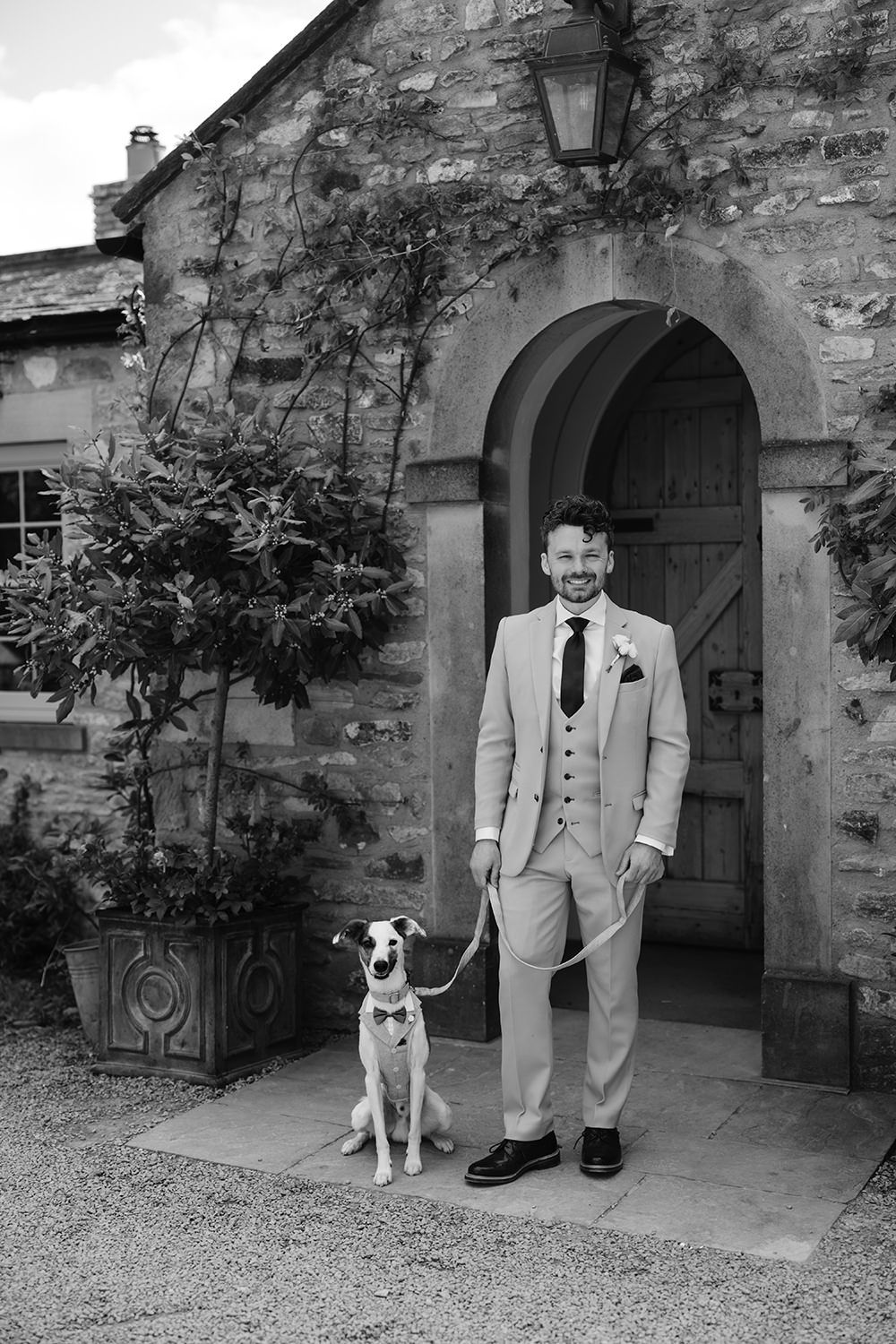 A black and white photo showing a man in a light colored suit standing beside his dog at a traditional stone entryway.